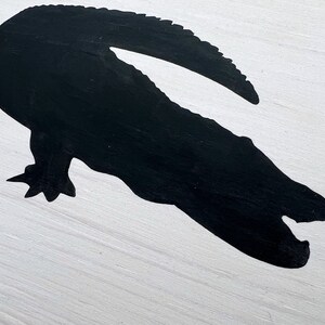 Puede incluir: Una silueta negra de un cocodrilo con la boca abierta, pintada sobre una superficie de madera blanca.
