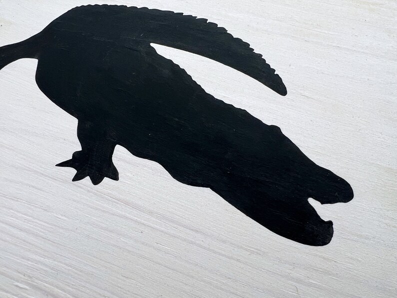 Puede incluir: Una silueta negra de un cocodrilo con la boca abierta, pintada sobre una superficie de madera blanca.
