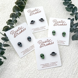 May include: Four pairs of alien-themed stud earrings on white cards with the text "Rustic Strokes". The earrings feature gray alien heads and gray UFOs, and green alien heads. The cards also include text about being hypoallergenic and handmade in Florida.