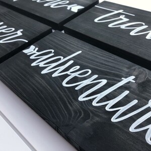 May include: Set of five black wooden signs with white calligraphy. The signs read "discover", "adventure", "travel", "explore", and "wander". Each sign has a small white arrow pointing to the right.