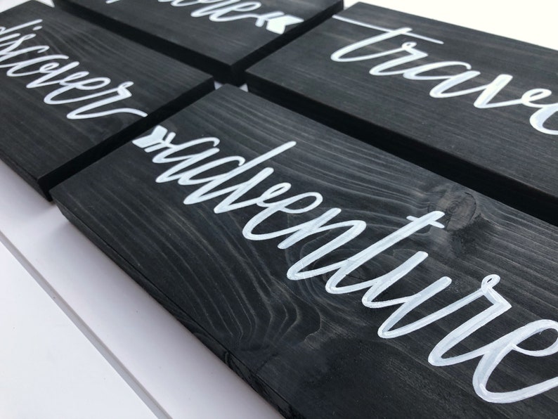 May include: Set of five black wooden signs with white calligraphy. The signs read "discover", "adventure", "travel", "explore", and "wander". Each sign has a small white arrow pointing to the right.