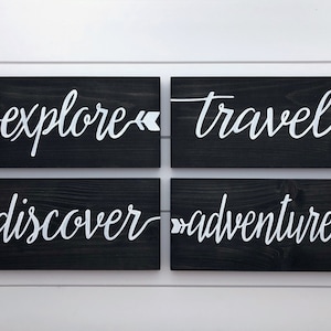 May include: Four black wooden signs with white lettering. The signs read "explore", "travel", "discover", and "adventure". Each sign has a white arrow pointing in the direction of the text.