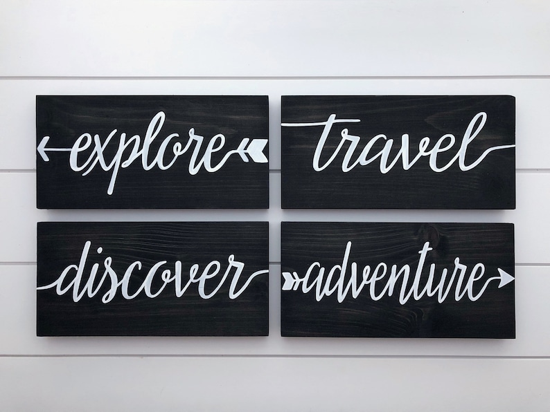 May include: Four black wooden signs with white lettering. The signs read "explore", "travel", "discover", and "adventure". Each sign has a white arrow pointing in the direction of the text.