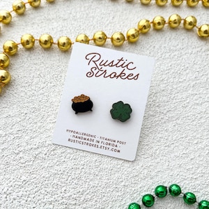 May include: A pair of wooden earrings featuring a pot of gold and a four-leaf clover. The earrings are hypoallergenic and have titanium posts. The earrings are handmade in Florida by Rustic Strokes.