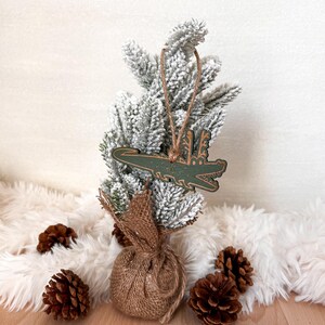 Puede incluir: Un adorno navide&ntilde;o verde con forma de cocodrilo cuelga de un peque&ntilde;o &aacute;rbol de Navidad cubierto de nieve. El &aacute;rbol est&aacute; sobre una superficie blanca y esponjosa, rodeado de pi&ntilde;as y una base envuelta en arpillera.