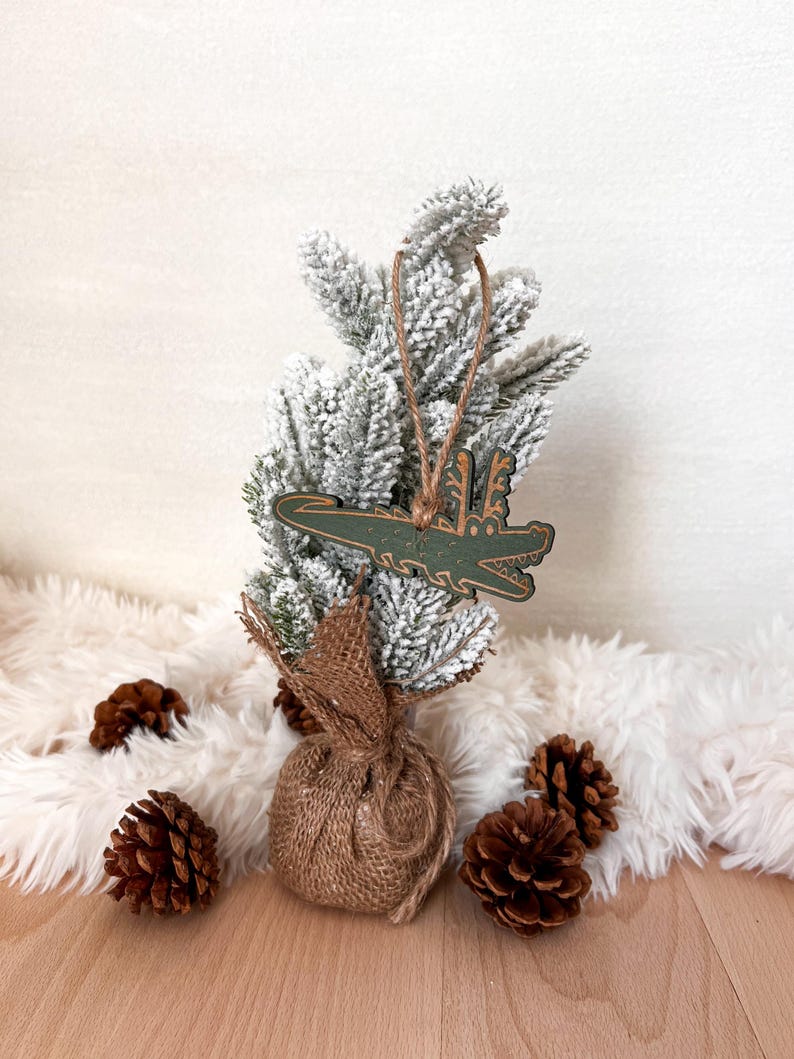 Puede incluir: Un adorno navide&ntilde;o verde con forma de cocodrilo cuelga de un peque&ntilde;o &aacute;rbol de Navidad cubierto de nieve. El &aacute;rbol est&aacute; sobre una superficie blanca y esponjosa, rodeado de pi&ntilde;as y una base envuelta en arpillera.