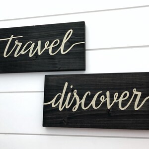 May include: Two wooden signs with the words "travel" and "discover" written in white cursive script on a black background.