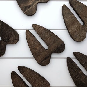 K&ouml;nnte beinhalten: Sechs dunkelbraune, aus Holz ausgeschnittene Formen, die an Hirschschenkel erinnern. Die Formen sind in einem Muster auf einer wei&szlig;en Oberfl&auml;che angeordnet.