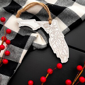 May include: A wooden Florida state ornament with floral detailing, hanging by a twine string. The ornament is set against a black and white checkered cloth and a black surface, with red berries in the foreground.