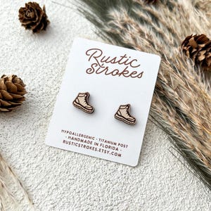 May include: Wooden stud earrings in the shape of hiking boots on a white card with the text "Rustic Strokes". The earrings are light brown with dark brown outlines. The card also includes text about the earrings being hypoallergenic and handmade.