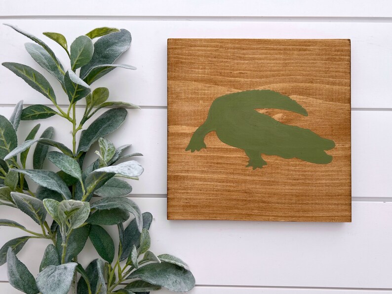 Puede incluir: Un letrero de madera con una silueta de cocodrilo verde pintada en &eacute;l. El letrero tiene un acabado marr&oacute;n r&uacute;stico.