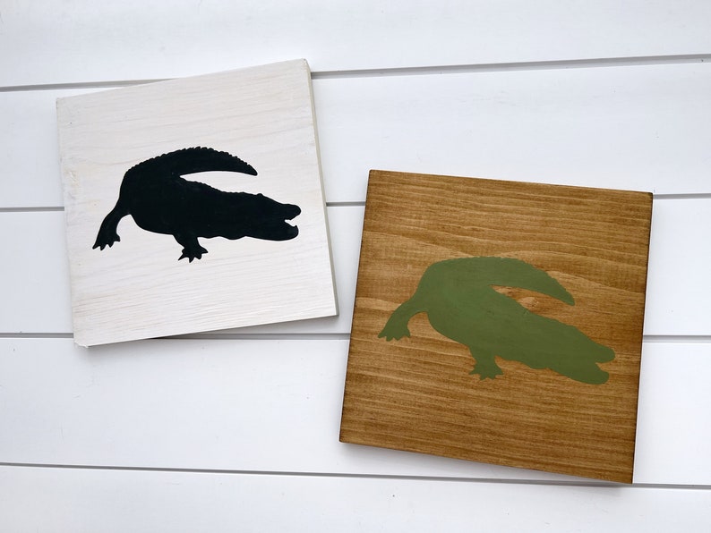Puede incluir: Dos letreros de madera con siluetas de caimanes. Un letrero es blanco con un caim&aacute;n negro, el otro es marr&oacute;n con un caim&aacute;n verde.