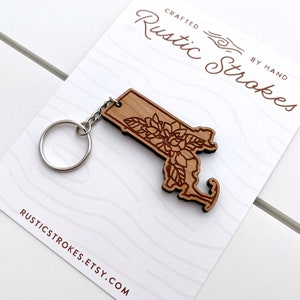 May include: A wooden keychain shaped like the state of Massachusetts with a floral design. The keychain is on a white card with the text "Crafted Rustic Strokes By Hand" and "rusticstrokes.etsy.com".