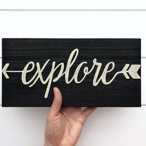 May include: A black wooden sign with the word "explore" written in white cursive script with arrows on either side of the word.