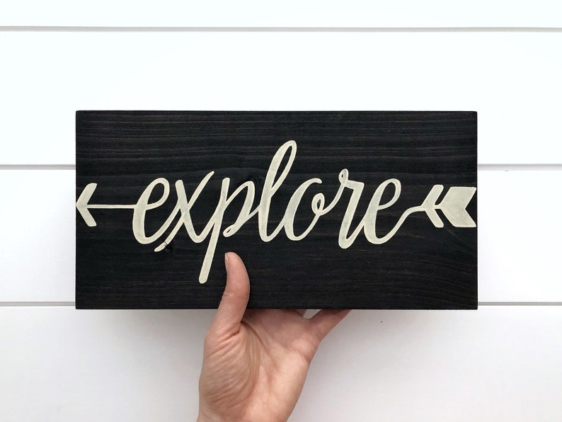 May include: A black wooden sign with the word "explore" written in white cursive script with arrows on either side of the word.