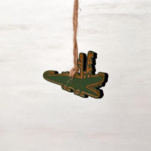 Puede incluir: Adorno de madera de un cocodrilo verde con detalles dorados, colgado de una cuerda de hilo natural. El cocodrilo tiene la boca abierta y detalles decorativos. El fondo es de color claro y neutro.