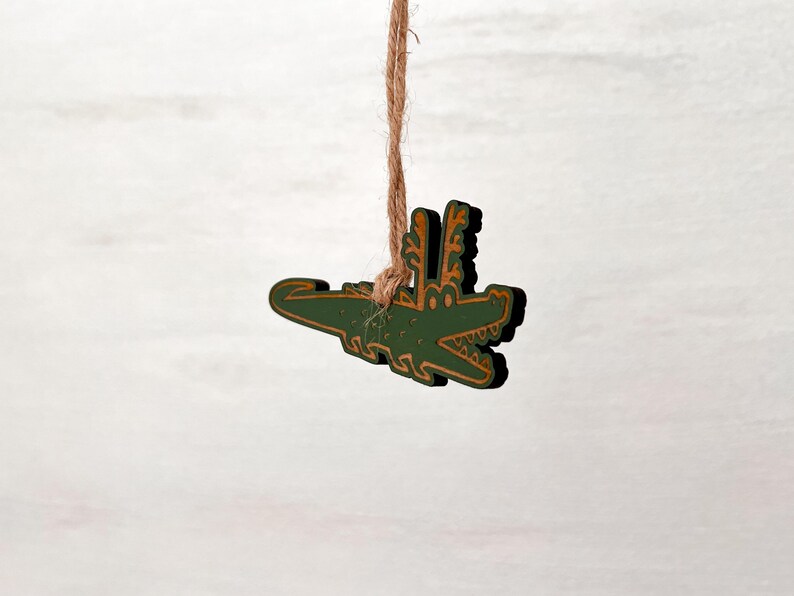 Puede incluir: Adorno de madera de un cocodrilo verde con detalles dorados, colgado de una cuerda de hilo natural. El cocodrilo tiene la boca abierta y detalles decorativos. El fondo es de color claro y neutro.