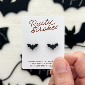 May include: A pair of black bat-shaped stud earrings on a white card with the text "Rustic Strokes". The card also includes the text "Hypoallergenic - Titanium - Handmade in Florida - Rusticstrokes.etsy.com".