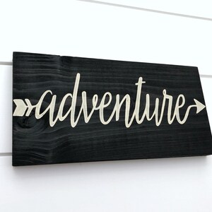 May include: Black wooden sign with the word "adventure" written in white script with an arrow pointing to the right.
