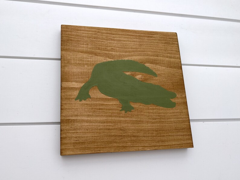 Puede incluir: Un letrero de madera con una silueta de cocodrilo verde pintada en &eacute;l.