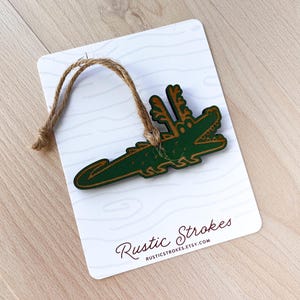 Puede incluir: Adorno de madera con forma de cocodrilo verde con detalles marrones y una cuerda de yute para colgar. El adorno est&aacute; sujeto a una tarjeta blanca con un patr&oacute;n de vetas de madera y el texto "Rustic Strokes".