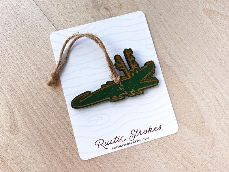 Puede incluir: Adorno de madera con forma de cocodrilo verde con detalles marrones y una cuerda de yute para colgar. El adorno est&aacute; sujeto a una tarjeta blanca con un patr&oacute;n de vetas de madera y el texto "Rustic Strokes".