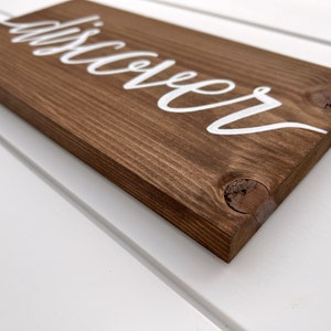 May include: A wooden sign with the word "discover" painted in white cursive lettering.