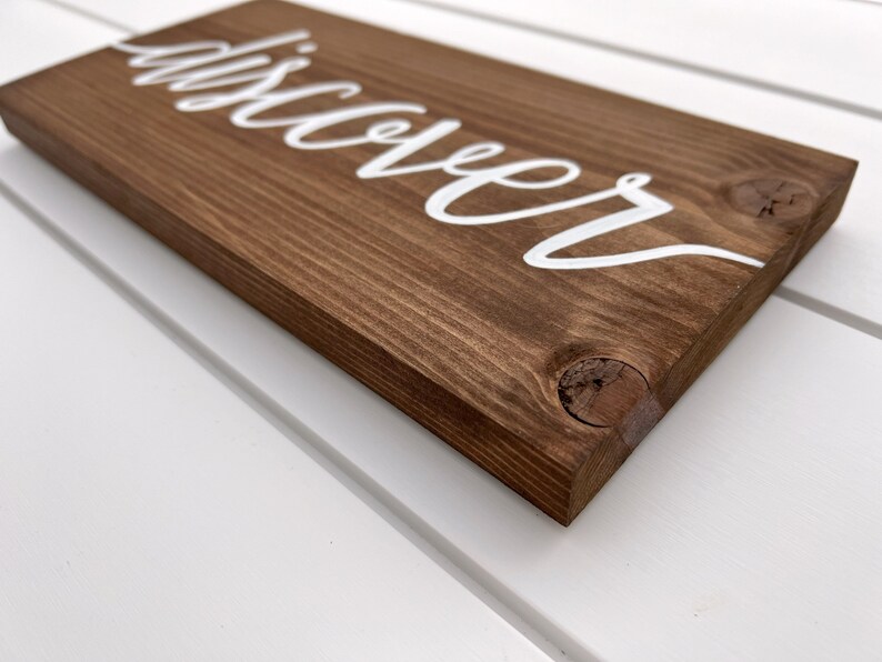 May include: A wooden sign with the word "discover" painted in white cursive lettering.
