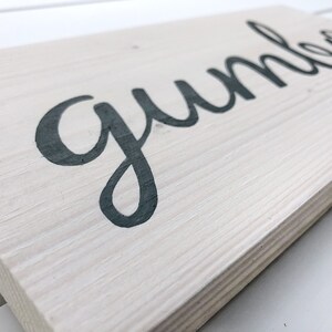 May include: A rectangular wooden sign with the word "gumbo" in dark grey cursive lettering. The sign has a light grey, weathered finish, and the wood grain is visible. The sign is angled slightly, showing the texture of the wood.