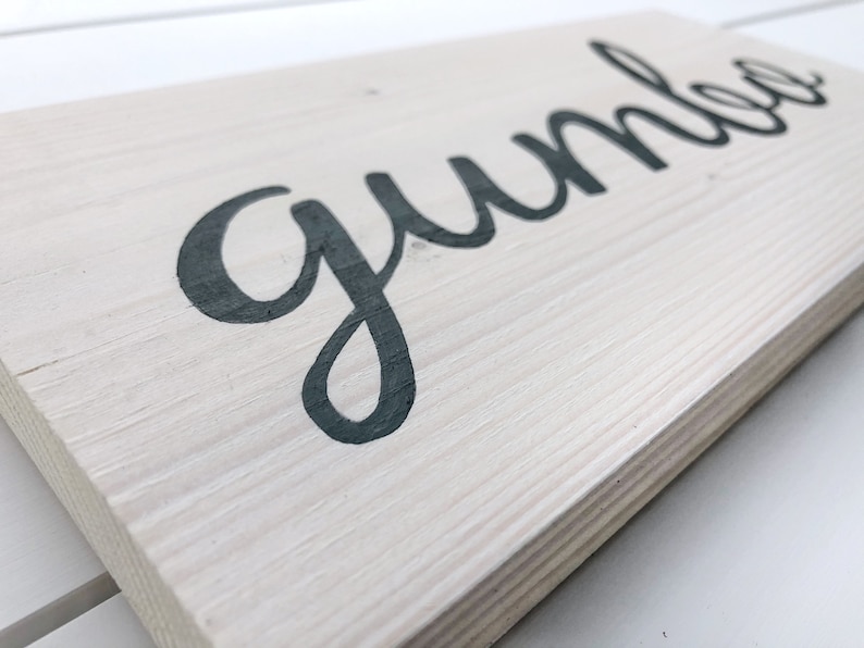 May include: A rectangular wooden sign with the word "gumbo" in dark grey cursive lettering. The sign has a light grey, weathered finish, and the wood grain is visible. The sign is angled slightly, showing the texture of the wood.