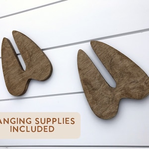 K&ouml;nnte beinhalten: Zwei braune Holz-Tierabdr&uuml;cke, die an einer wei&szlig;en Holzwand befestigt sind. Der Text "HANGING SUPPLIES INCLUDED" ist auf einem beigefarbenen Rechteck unter den Abdr&uuml;cken gedruckt.