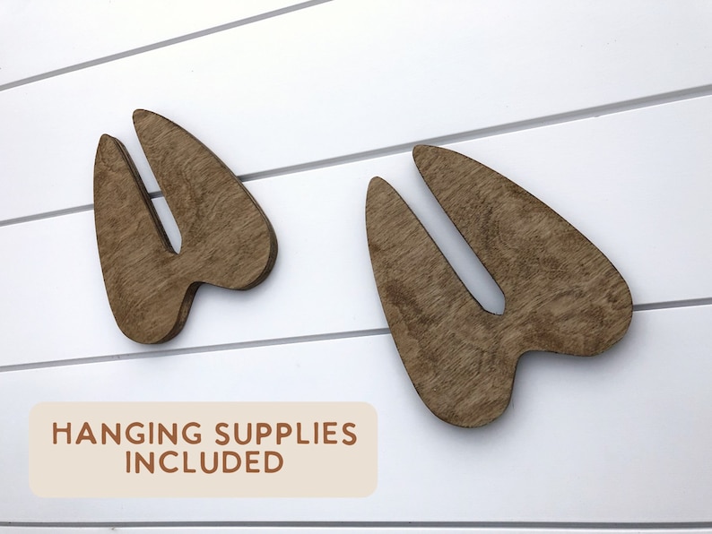 K&ouml;nnte beinhalten: Zwei braune Holz-Tierabdr&uuml;cke, die an einer wei&szlig;en Holzwand befestigt sind. Der Text "HANGING SUPPLIES INCLUDED" ist auf einem beigefarbenen Rechteck unter den Abdr&uuml;cken gedruckt.