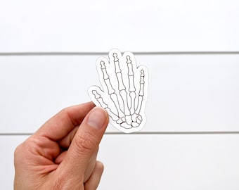 May include: A white sticker featuring a detailed outline of a human hand skeleton. The skeletal hand is depicted with visible bones and joints, perfect for Halloween or medical themes.