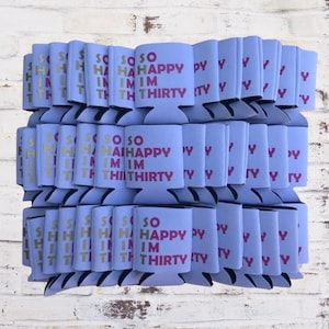 May include: A stack of light blue can coolers with the text "SO HAPPY I'M THIRTY" in pink and gold lettering. The coolers are made of a foam-like material and are designed to keep beverages cold. The background is a white brick wall.