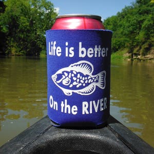 May include: A blue can cooler with the text "Life is better On the RIVER" and a white fish graphic. The cooler is wrapped around a red can, with a river and trees in the background.