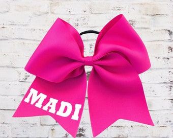 May include: A large pink cheer bow with a white name "MADI" on the front. The bow is tied with a black elastic band.