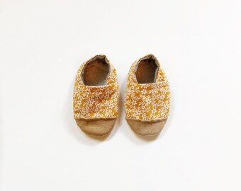 Può includere: Un paio di scarpine per neonati in tessuto giallo con un motivo floreale bianco. Le scarpine hanno una suola in camoscio marrone.