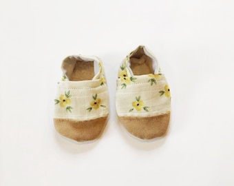 Può includere: Un paio di scarpine per neonati con tomaia in tessuto beige e suola in camoscio marrone. Le scarpine hanno un motivo floreale giallo.