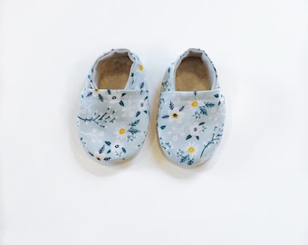Può includere: Un paio di scarpine per neonati di colore azzurro chiaro con un motivo floreale. Le scarpine hanno uno sfondo bianco con piccoli fiori gialli e foglie verdi.