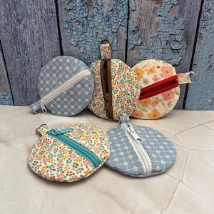 Könnte beinhalten: Fünf kleine, runde Münztaschen. Zwei sind hellblau mit weißem Blumenmuster und weißen Reißverschlüssen. Die anderen haben Blumenmuster mit türkisfarbenen und braunen Reißverschlüssen. Eine hat einen roten Reißverschluss.