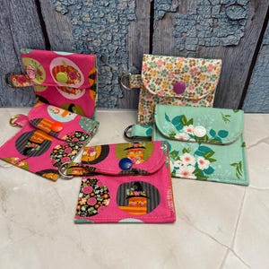 May include: A collection of small, fabric coin purses in various floral and patterned designs. The purses feature a snap closure and a metal ring for attachment. Colors include pink, green, and white.