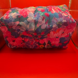 May include: A rectangular quilted pouch featuring a vibrant floral pattern with shades of hot pink, magenta, deep blue, and teal. The fabric is stitched with vertical lines, creating a soft, padded texture across the entire surface of the bag.