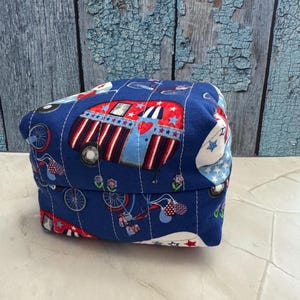 May include: A blue fabric box with a quilted design featuring red, white, and blue camper vans, stars, and bicycles. The box is likely used for storage or as a decorative item, with a patriotic theme.