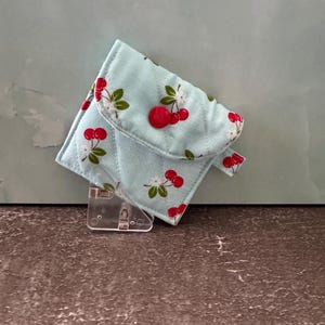 May include: A small, quilted wallet in a light blue fabric with a cherry pattern. The wallet has a flap secured by a red button. The cherry design features red cherries, white flowers, and green leaves. The wallet is displayed on a clear plastic stand.