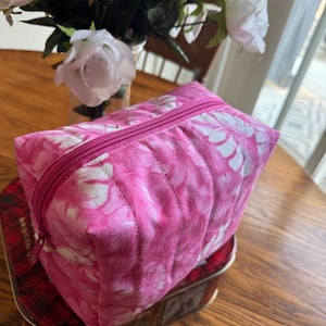 May include: A quilted, rectangular cosmetic bag in shades of pink with a white floral pattern. The bag has a matching pink zipper and is sitting on a red plaid tin. White roses are in the background.