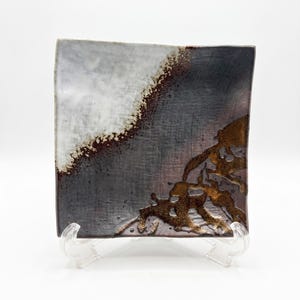 May include: A square ceramic plate featuring a textured surface with a color gradient transitioning from white to dark gray. The plate displays a distinct diagonal band of speckled brown and metallic bronze glaze accents along the edge and corner.