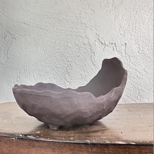May include: A handcrafted, dark gray ceramic bowl with a unique, wavy rim. The bowl rests on three small feet and has a textured surface. This decorative piece is suitable for various uses.