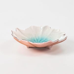 May include: A decorative ceramic bowl in the shape of a flower. The bowl has a white interior with a blue center, a pink exterior, and silver trim. It rests on three small feet.