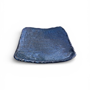 May include: A square, handmade ceramic plate with a dark blue glaze. The plate features a textured surface with concentric circular patterns and a slightly irregular edge. The plate is approximately 8 inches wide.