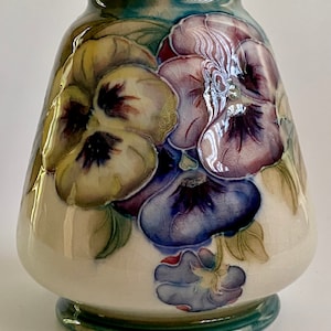 May include: A white ceramic vase with a blue, green, and purple floral design. The vase features a cluster of pansies with yellow, purple, and white petals.
